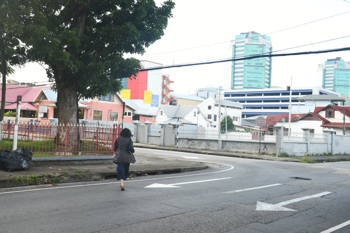 A woman walks along a curved road corner in an urban setting, with a large tree on the left, low-rise buildings, and two tall modern buildings in the background under an overcast sky.
