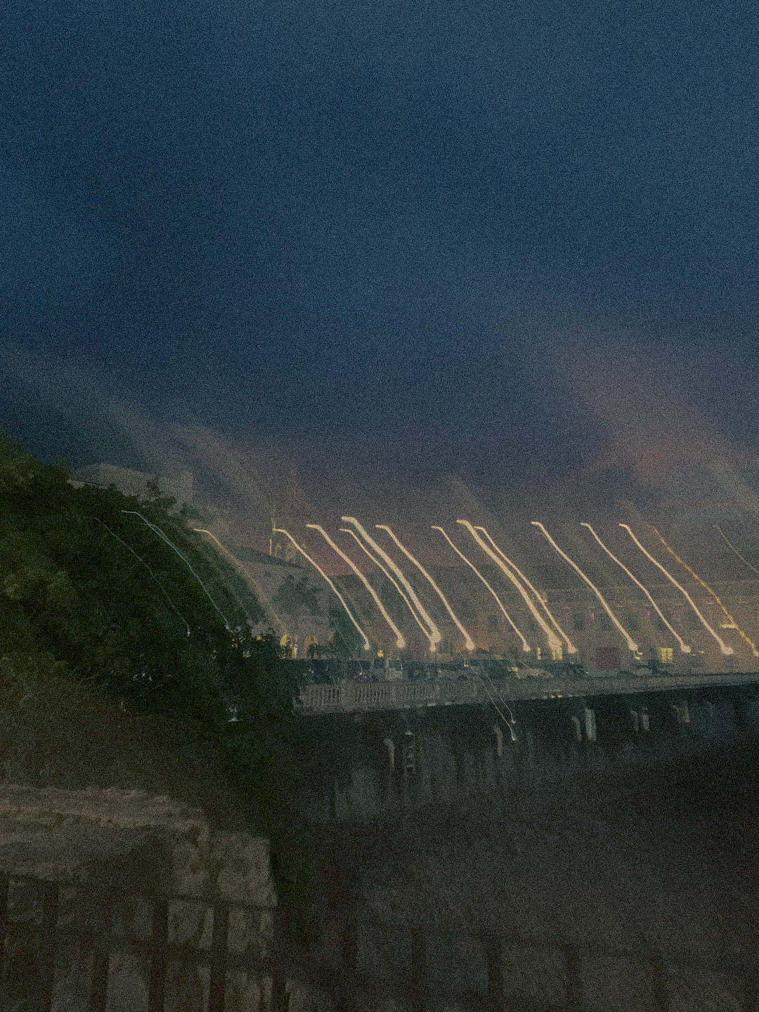 A grainy, atmospheric photograph of Panama City at night, with the illuminated arches of the dam and water jets against a dark blue sky.