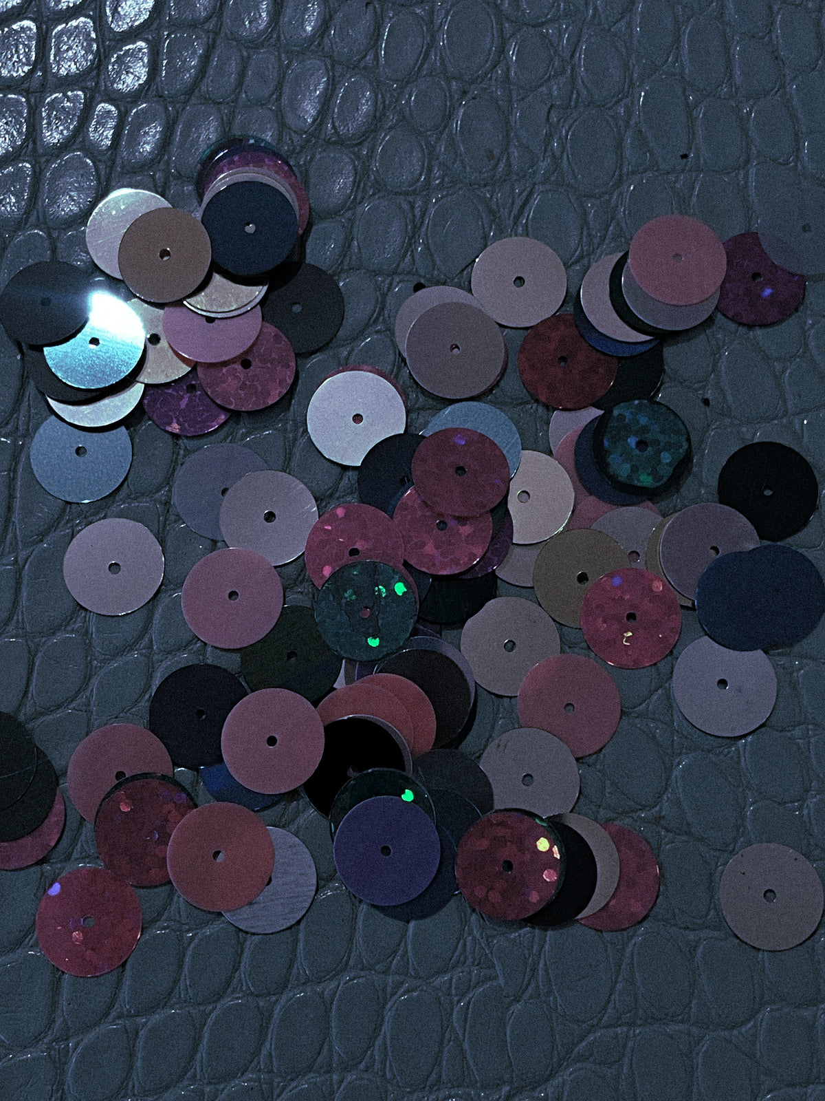 A scattered pile of round sequins in assorted colors including pink, purple, black, silver, and glittery shades, placed on a dark textured crocodile-embossed surface.