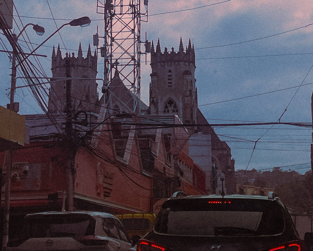 A low-angle street scene at dusk showing a church with two towers and a tall metal antenna structure in the center, surrounded by buildings, overhead wires, and cars in the foreground under a blue and pink sky.