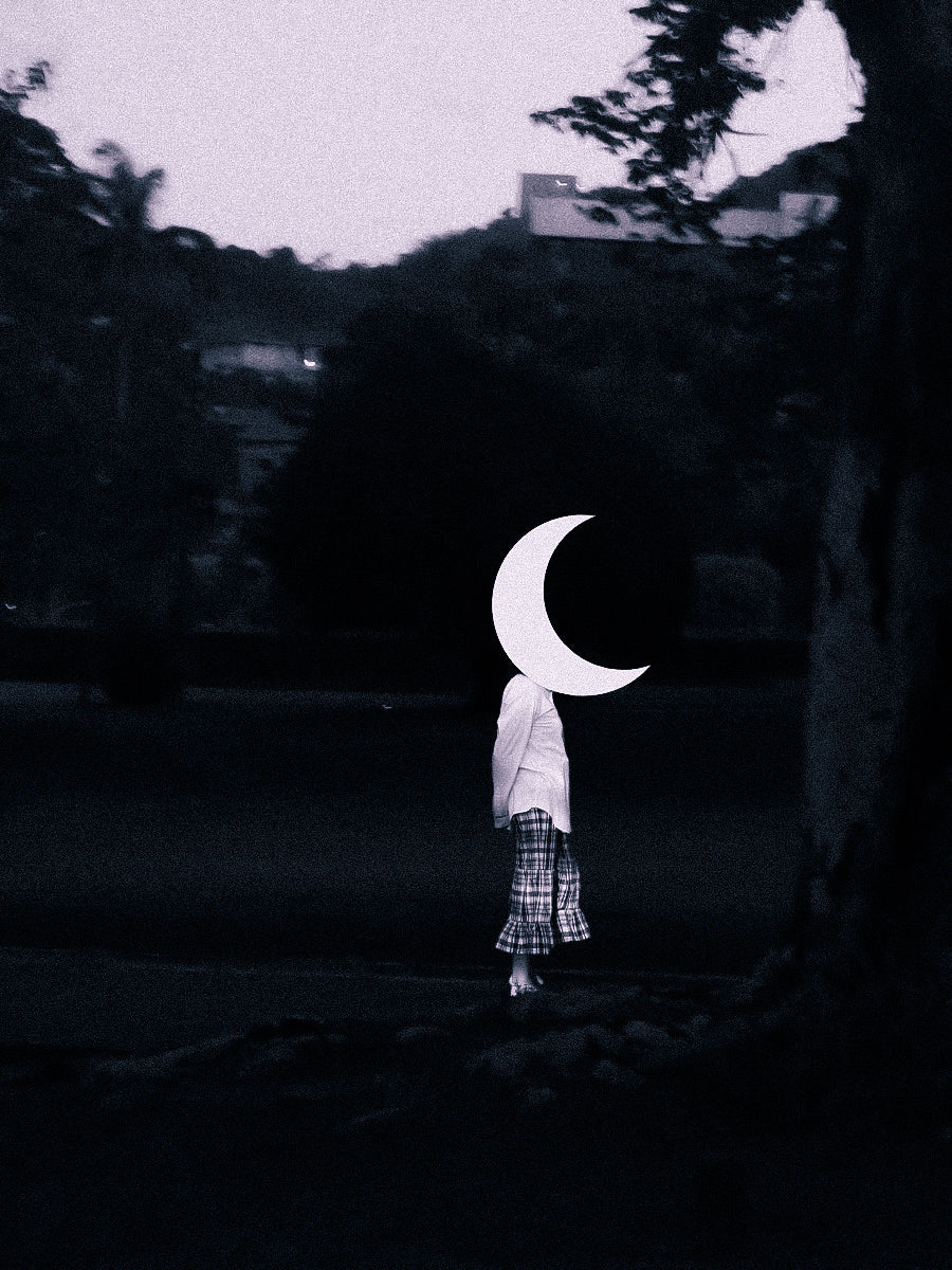 A person standing outdoors at dusk or dawn, wearing a white top and a black & white madras Àjẹ́ pedal pusher pants, with a large white crescent moon shape obscuring their head. The background is very dark with trees and hills.